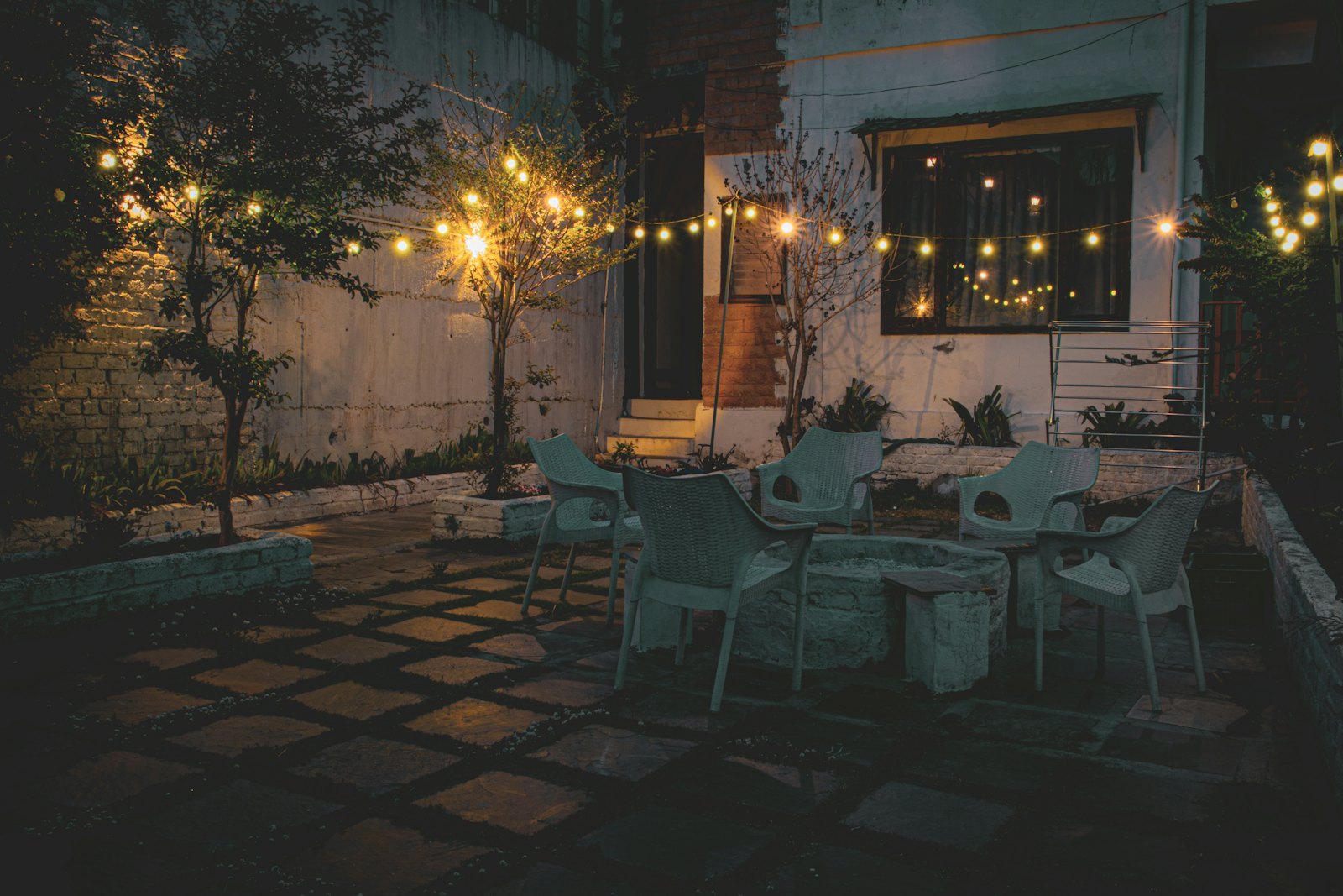 Paver patio at night under warm string lights, with chairs arranged around a round concrete centerpiece