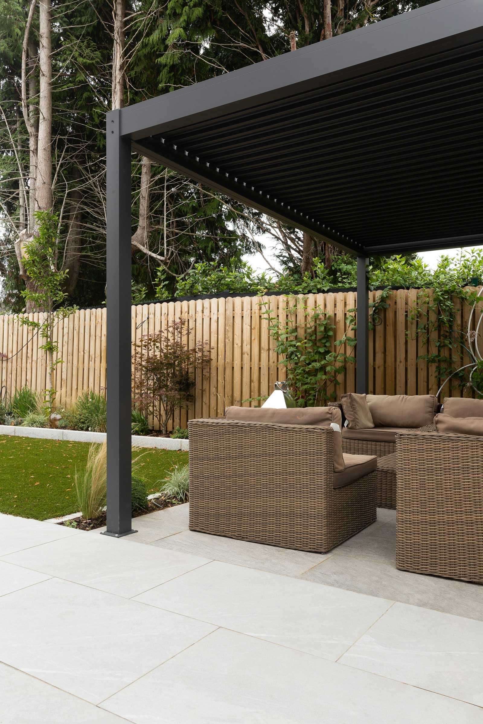 Wicker lounge set under a dark aluminum louvered pergola on a light paver patio with a wood privacy fence