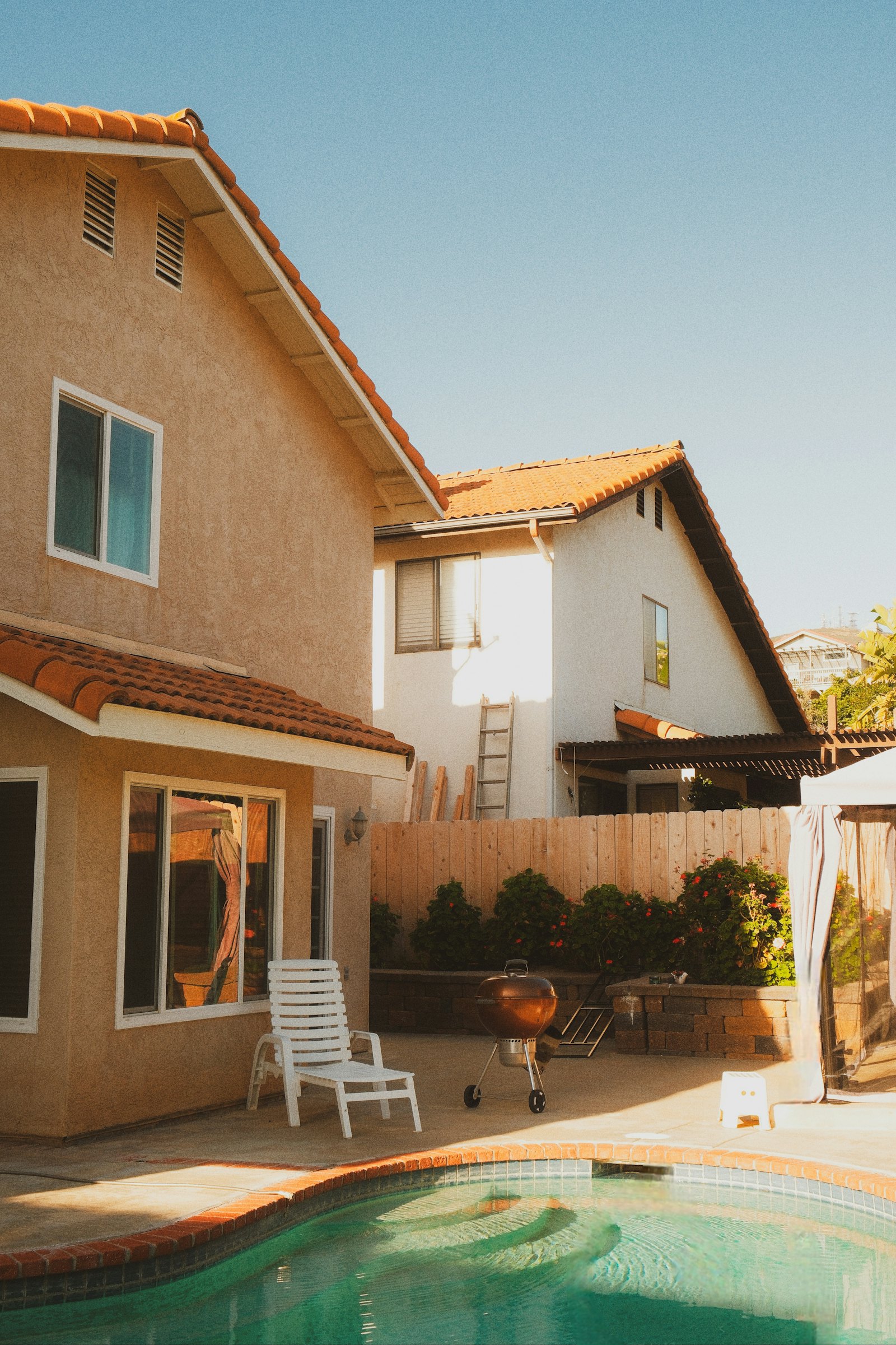 Backyard pool beside a two-story stucco home with a tile roof, wood fence and patio