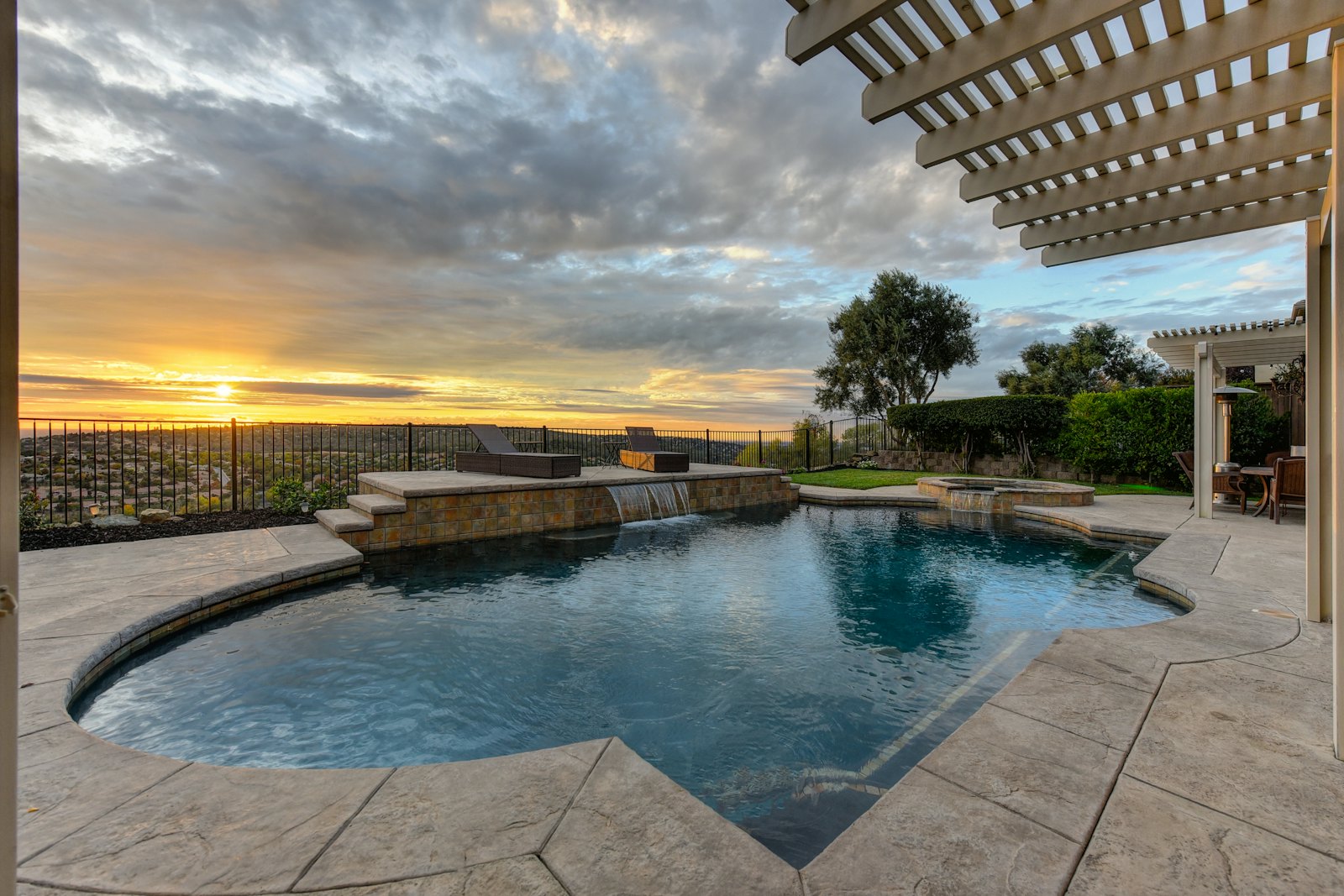 Freeform pool with a waterfall feature and a white pergola at sunset, on a patterned concrete deck